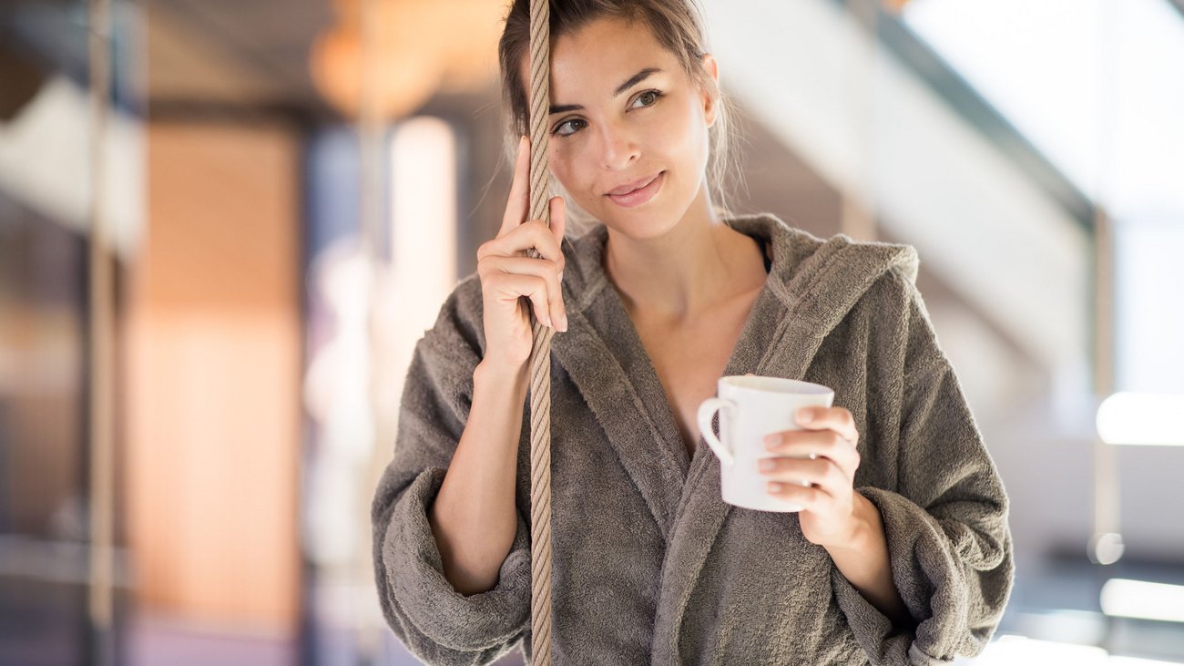 Frau in Bademantel hält Tasse und lehnt an Seil im Innenbereich