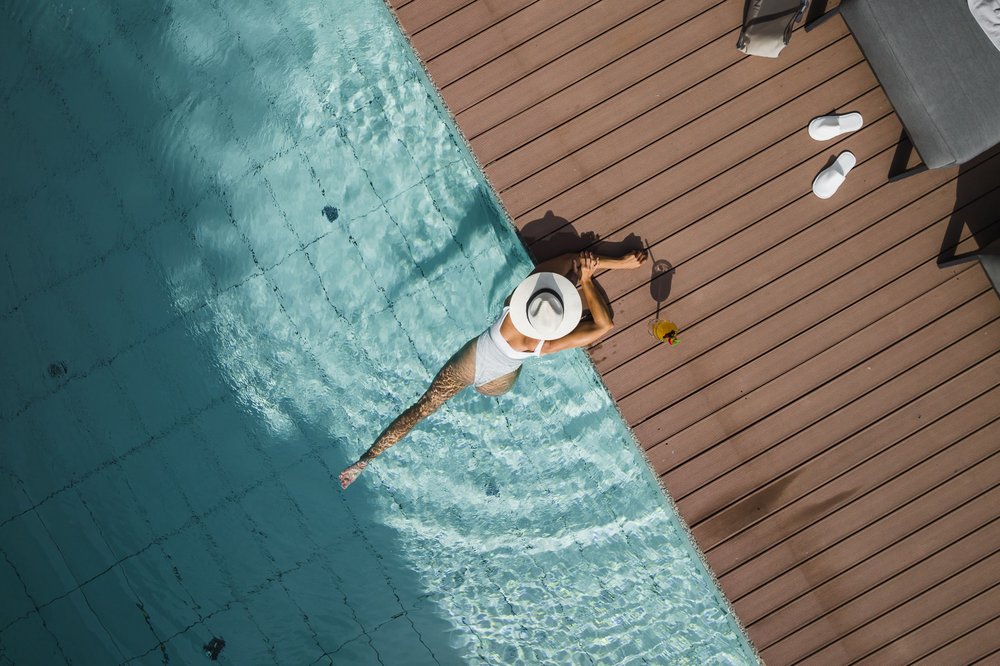 Frau entspannt am Poolrand in weißem Badeanzug und Hut mit Cocktail neben ihr