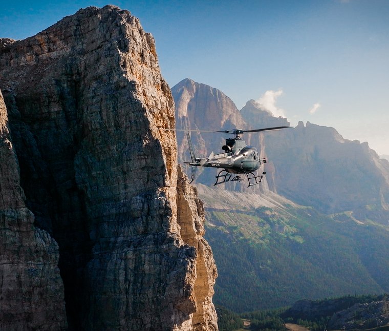 Winkler’s Stories: i nostri alberghi con maneggio di cavalli Elicottero vola accanto a una parete rocciosa ripida tra le montagne con cielo sereno