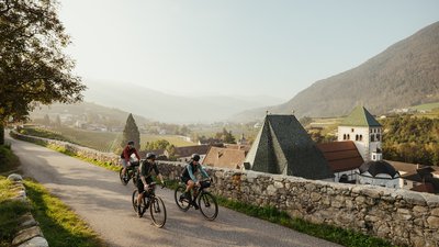 Tre ciclisti pedalano su una strada con panorama di montagna e villaggio al sole