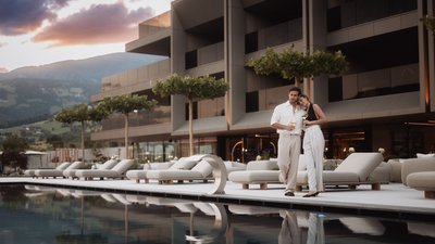 Paar mit Getränken am Pool vor modernem Gebäude bei Sonnenuntergang