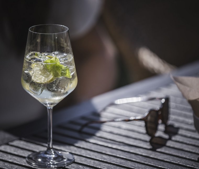 Winkler’s Stories: i nostri alberghi con maneggio di cavalli Bicchiere da vino con ghiaccio, menta e lime su un tavolo con occhiali da sole sullo sfondo