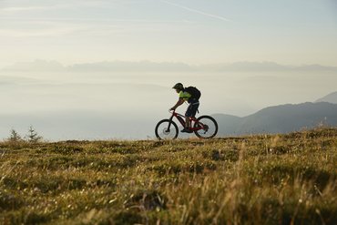 Mountainbiker fährt auf einem Grasweg mit nebelverhangenen Bergen im Hintergrund