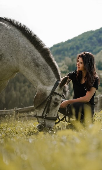 Donna accovacciata accanto a un cavallo che mangia erba in un prato montano