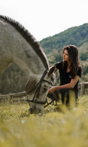 Woman kneeling beside a grazing horse in a meadow with mountains behind