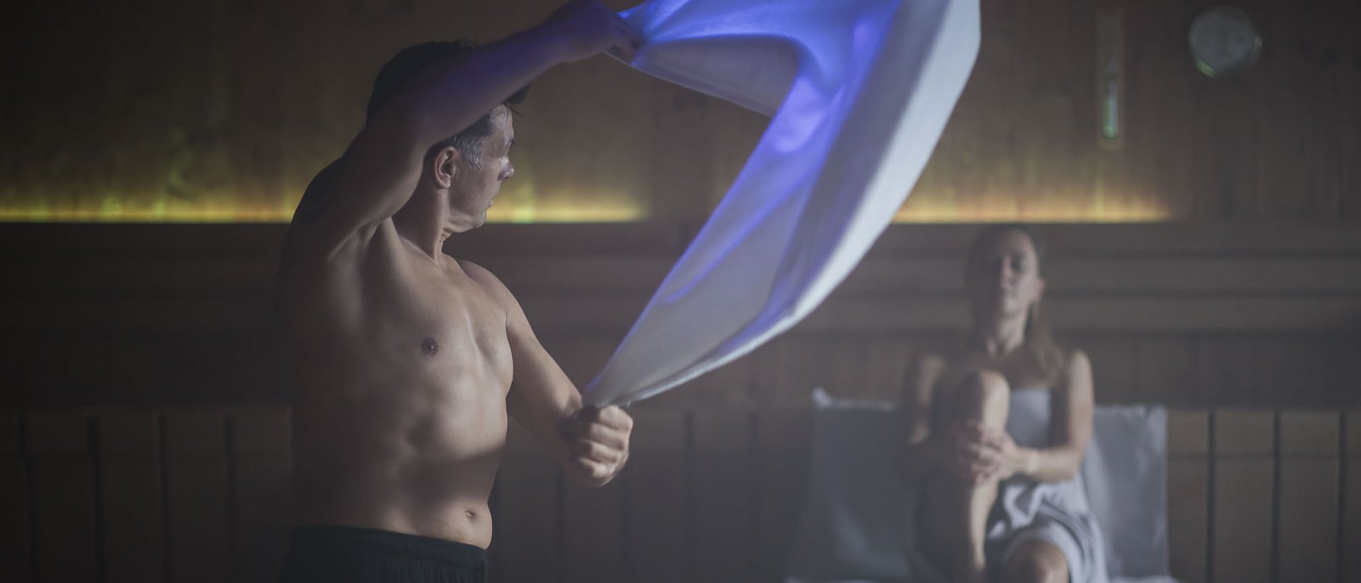 Mann schwingt Handtuch in Sauna, Frau sitzt entspannt auf Bank