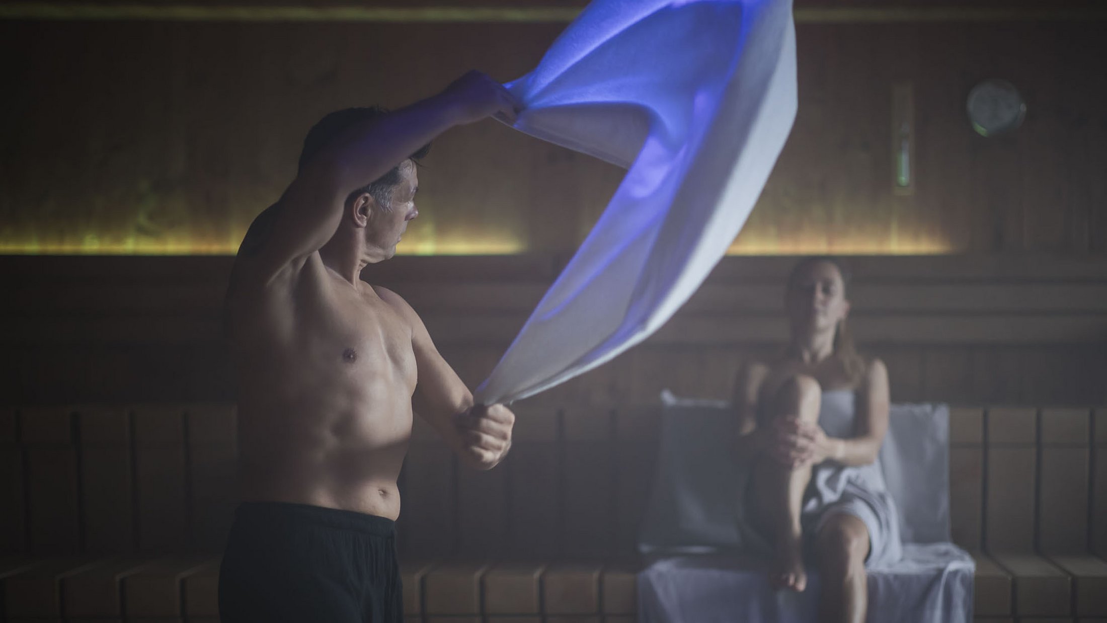 Mann schwingt Handtuch in Sauna, Frau sitzt entspannt auf Bank