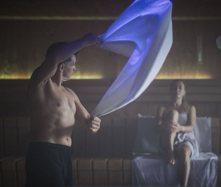 Mann schwingt Handtuch in Sauna, Frau sitzt entspannt auf Bank