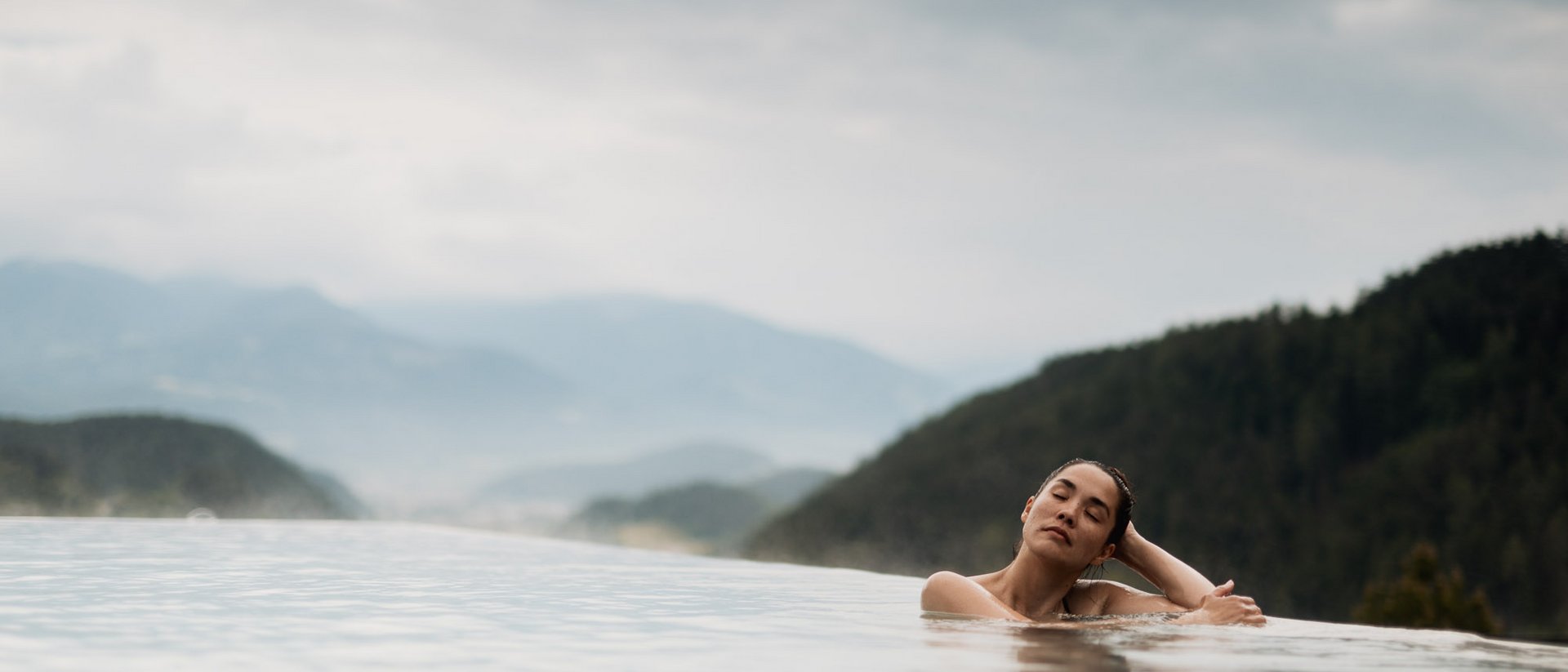 Woman relaxing in infinity pool with mountain views and cloudy sky