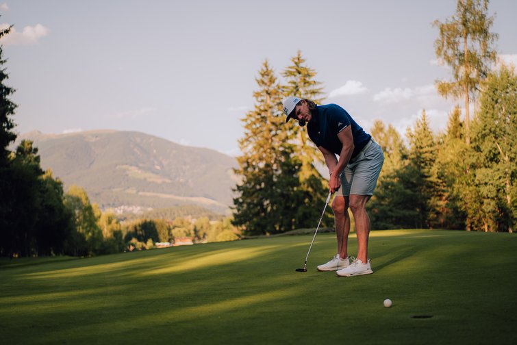 Platzreife in Südtirol erlangen: unser Premium-Golf-Service Golfspieler beim Putten auf einem Golfplatz vor bewaldeter Berglandschaft