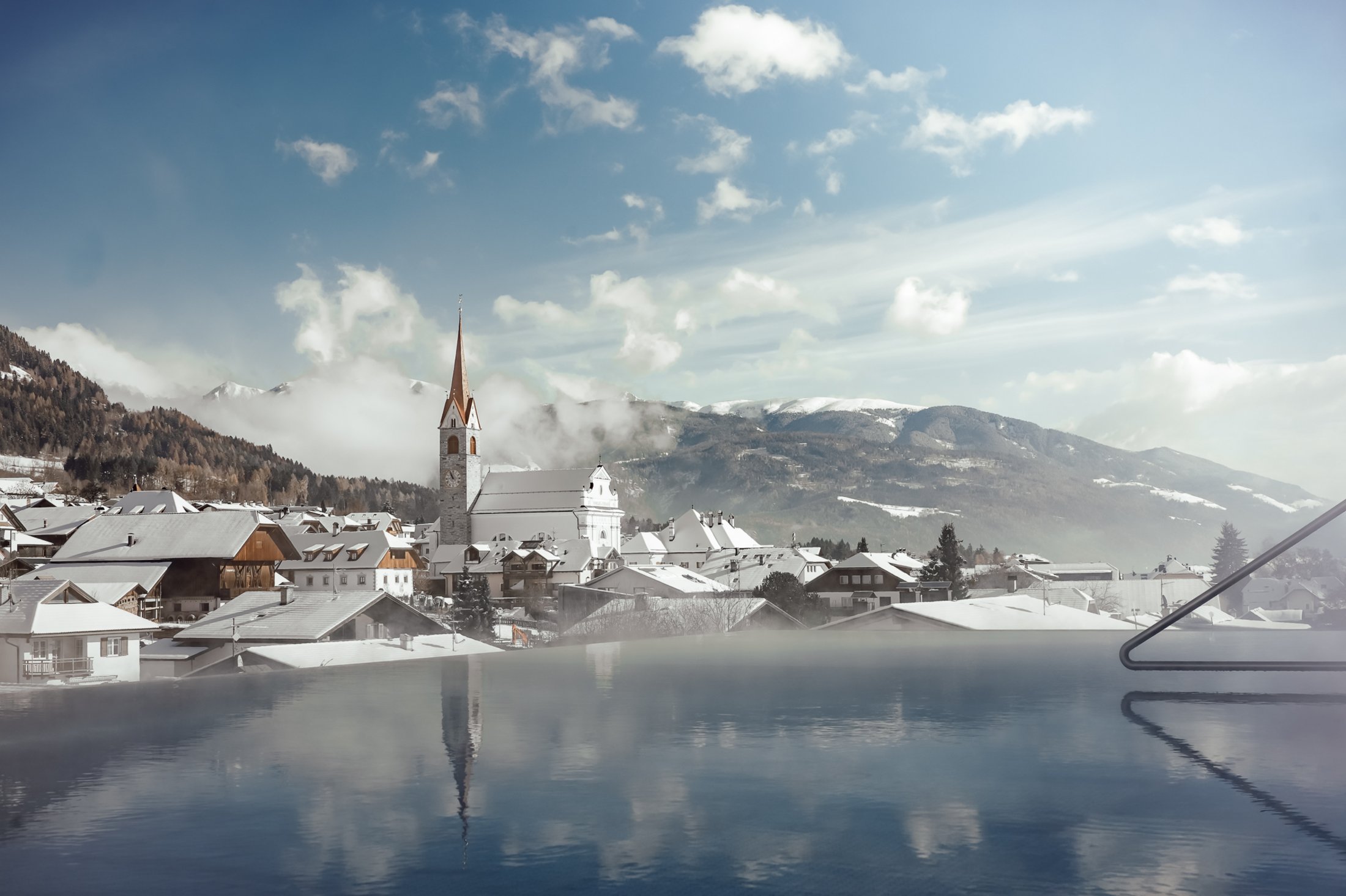 Die Heimat des Strahlens: Ihr Wellnesshotel in Südtirol Verschneites Dorf mit Kirche und Berglandschaft, Blick von einem Pool aus