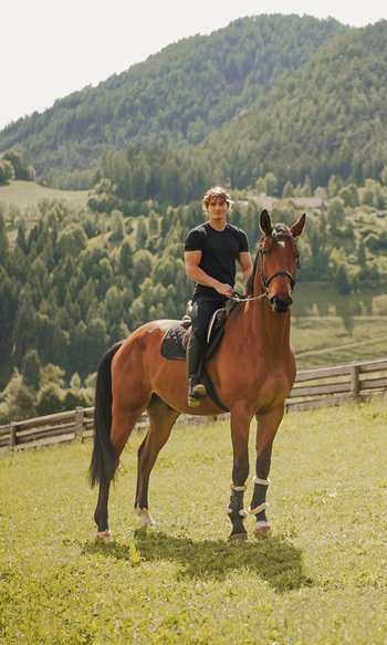 Mann reitet braunes Pferd auf einer Wiese mit Bergen im Hintergrund