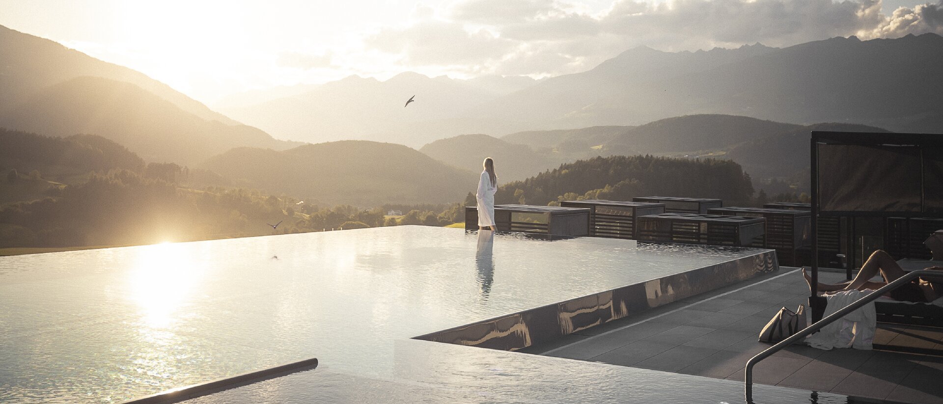 Your source of enrichment: our hotel at Mt Plan de Corones Woman in white robe at infinity pool with mountain view at sunset