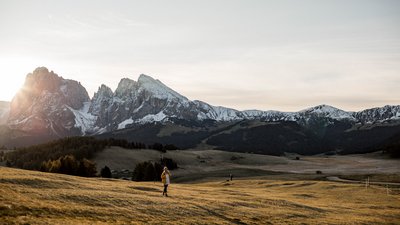 Donna cammina su un prato verde con montagne innevate al tramonto