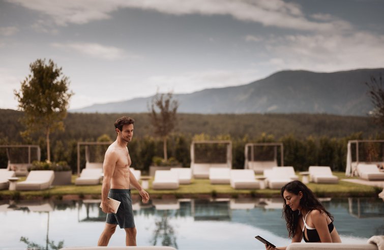 Mann in Badeshorts und Frau am Pool bei einem sonnigen Tag vor Bergkulisse