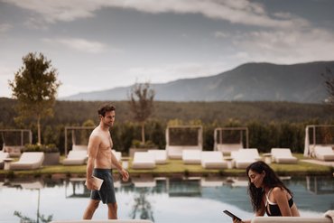 Mann in Badeshorts und Frau am Pool bei einem sonnigen Tag vor Bergkulisse