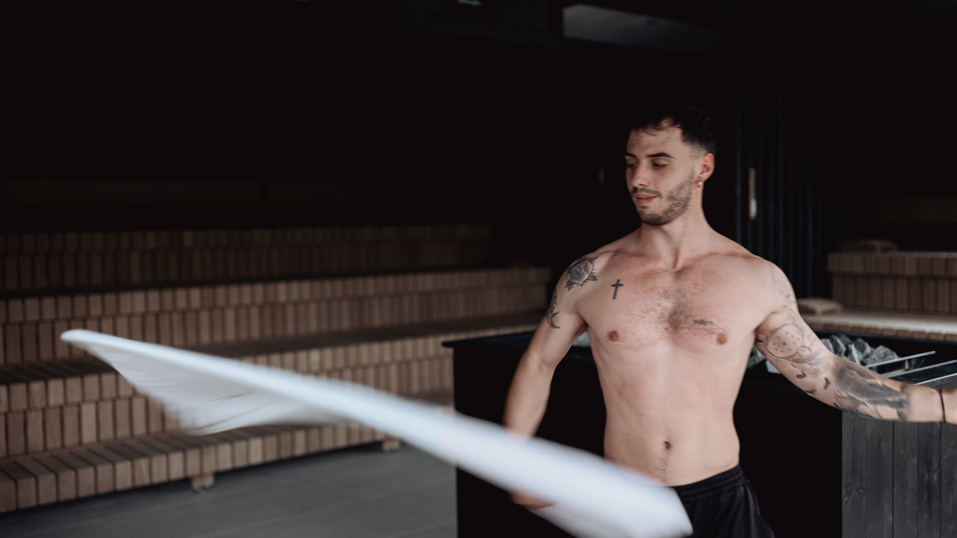 Oberkörperfreier Mann mit Tattoos winkt mit einem weißen Tuch in einer Sauna
