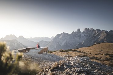 Zwei Wanderer sitzen auf Bank mit Blick auf Berglandschaft und blauen Himmel