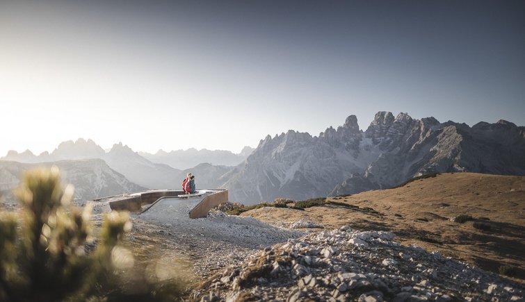Zwei Wanderer sitzen auf Bank mit Blick auf Berglandschaft und blauen Himmel