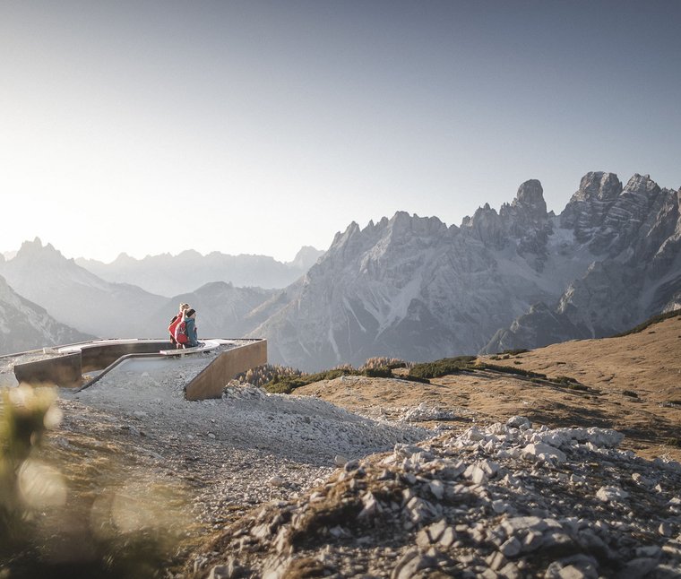 Due escursionisti seduti su panchina con vista sulle montagne e cielo blu