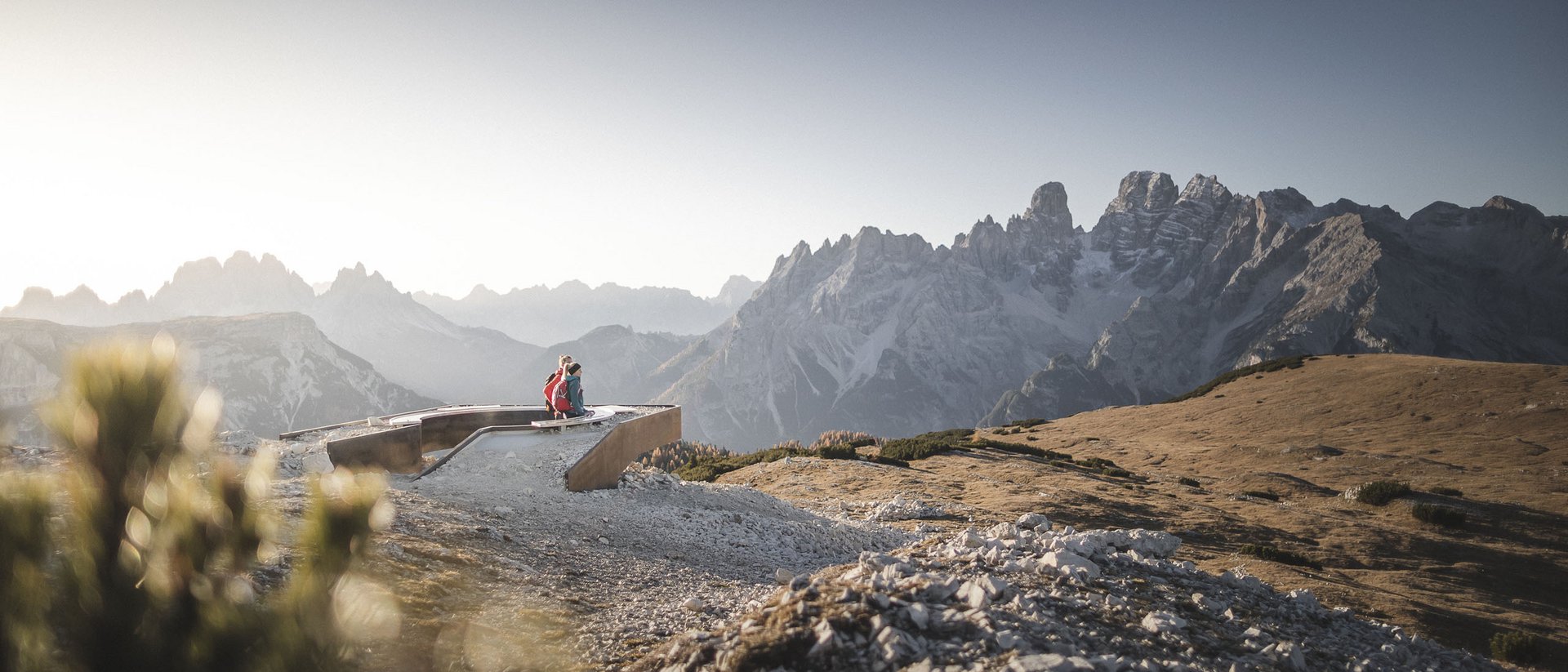 Zwei Wanderer sitzen auf Bank mit Blick auf Berglandschaft und blauen Himmel