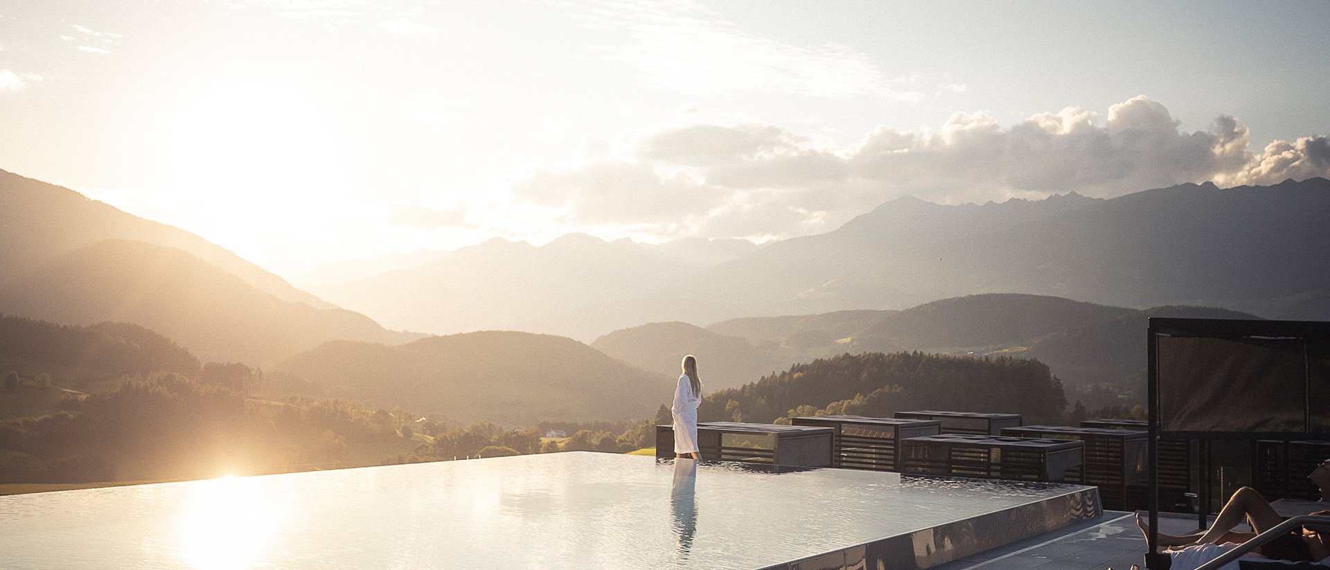 Frau im Bademantel am Infinity-Pool mit Bergpanorama bei Sonnenuntergang