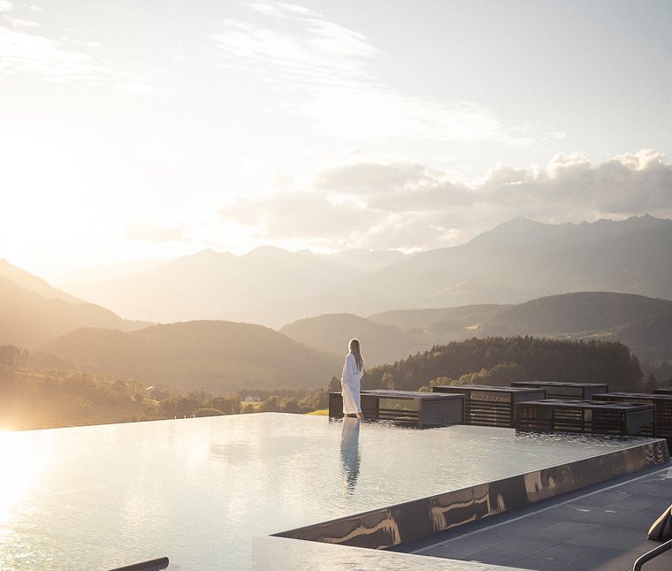 Eine regenerierende Auszeit vor Weihnachten Frau im Bademantel am Infinity-Pool mit Bergpanorama bei Sonnenuntergang