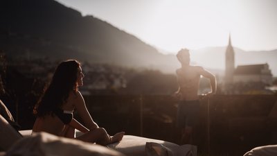 Photos from your hotel in Pfalzen in Val Pusteria/Pustertal Woman sitting on lounge chair and man leaning at sunset with mountain and church in background