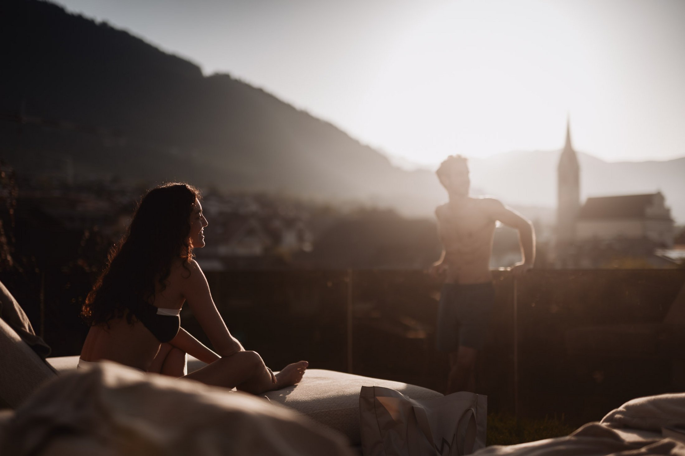 Donna seduta su lettino e uomo appoggiato al tramonto con montagna e chiesa sullo sfondo