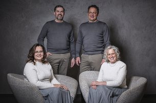 Two men standing behind two seated women, all smiling in neutral indoor setting