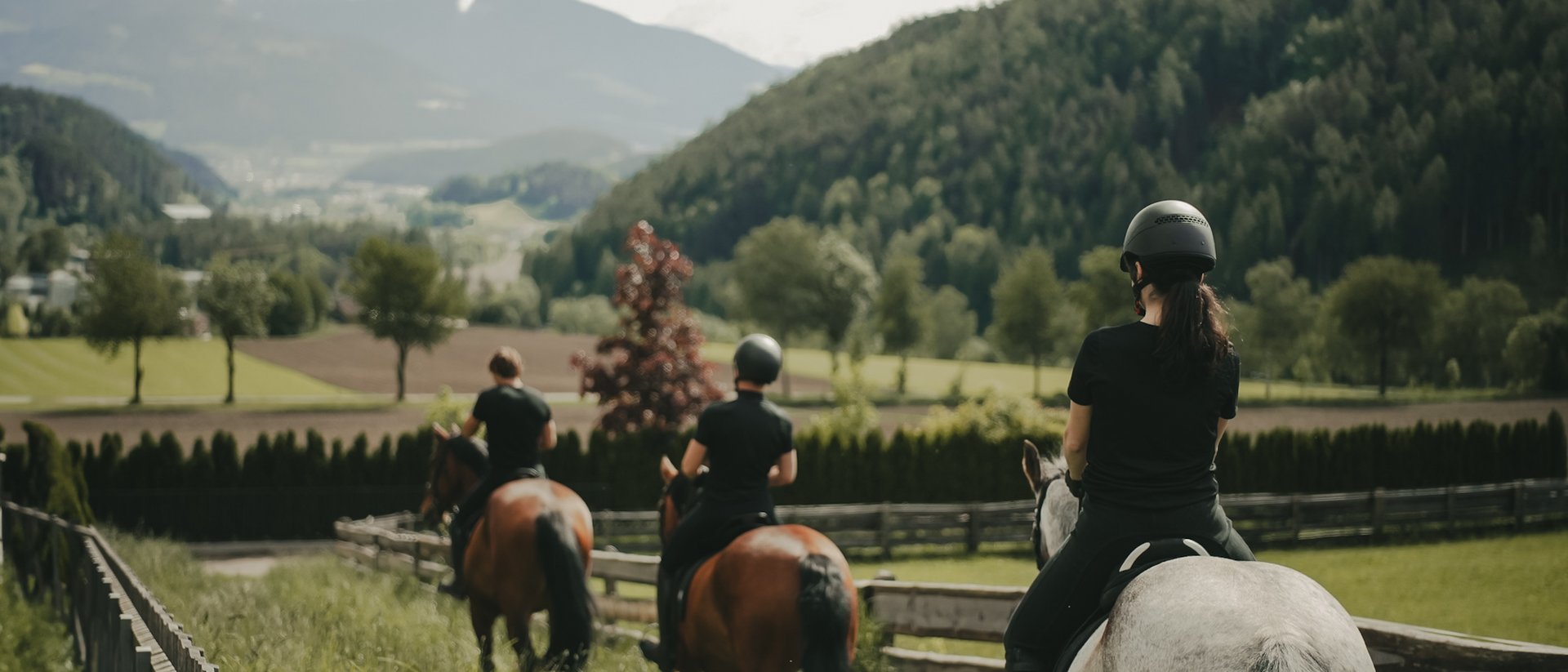 Dove fare equitazione in Alto Adige: hotel con maneggio Tre cavalieri a cavallo in un paesaggio verde con montagne sullo sfondo