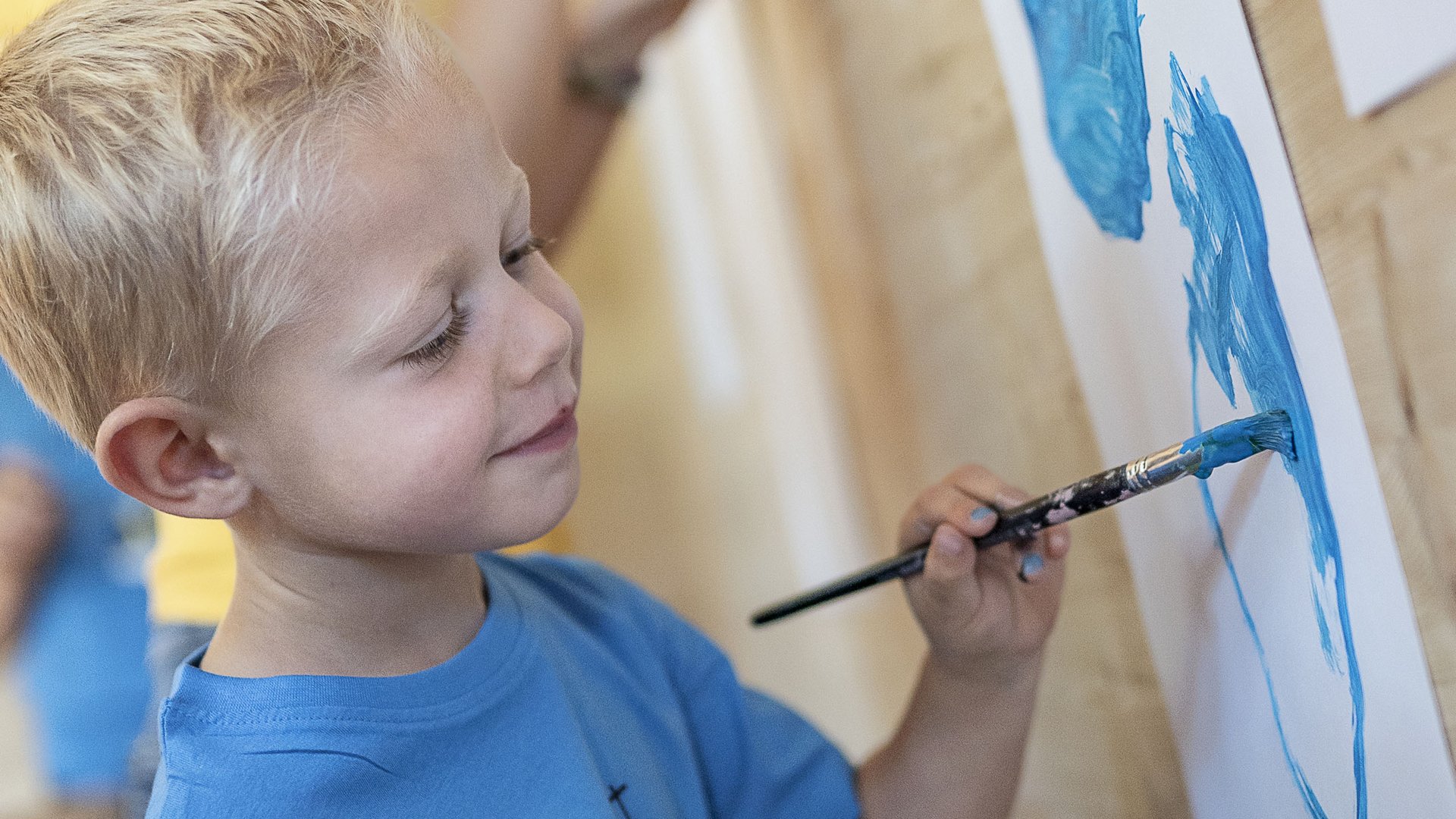 Child happily painting with blue paint on paper attached to the wall