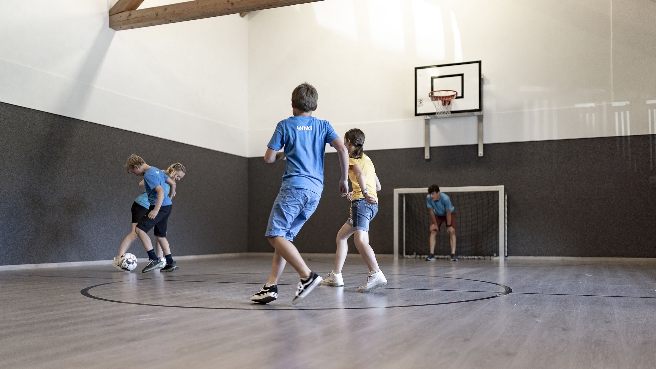 Children playing soccer in indoor gym with basketball hoop and goal