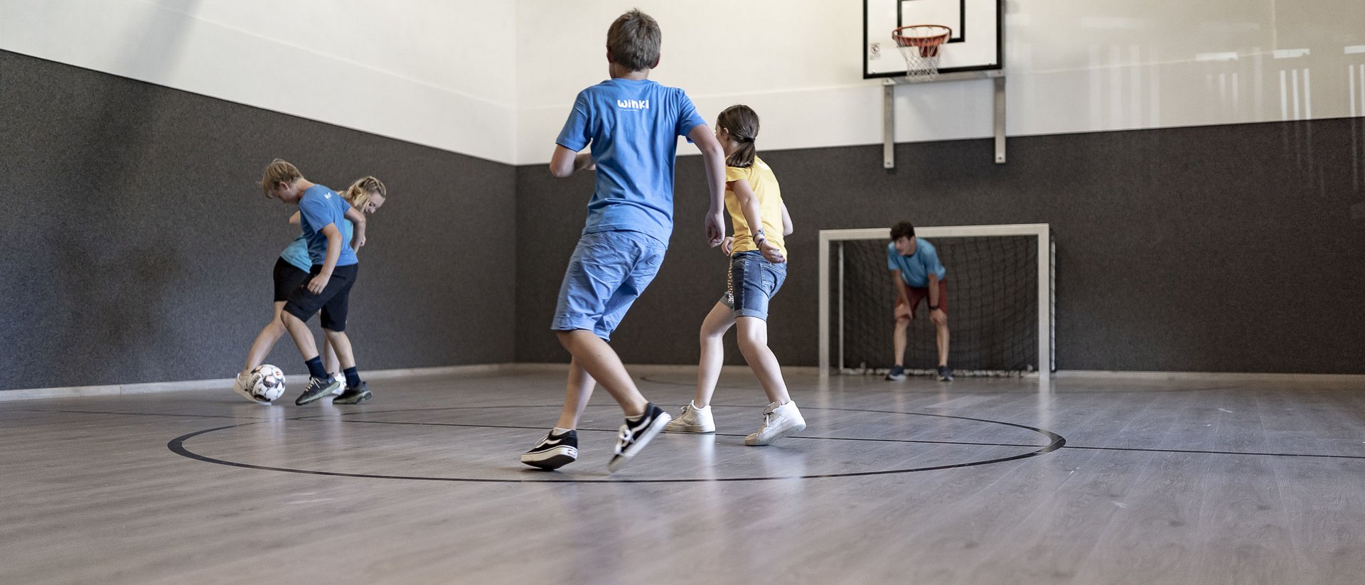 Children playing soccer in indoor gym with basketball hoop and goal