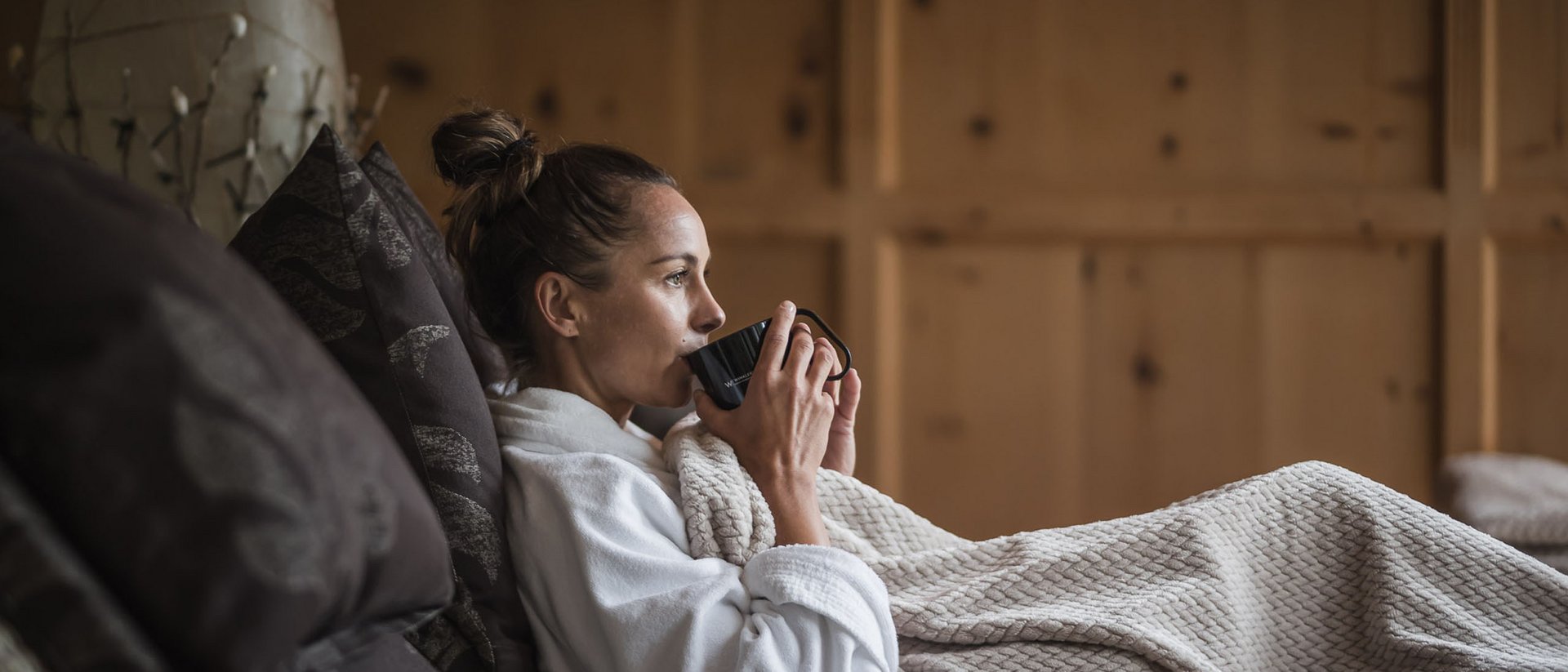 Donna in accappatoio che beve caffè su divano comodo con coperta