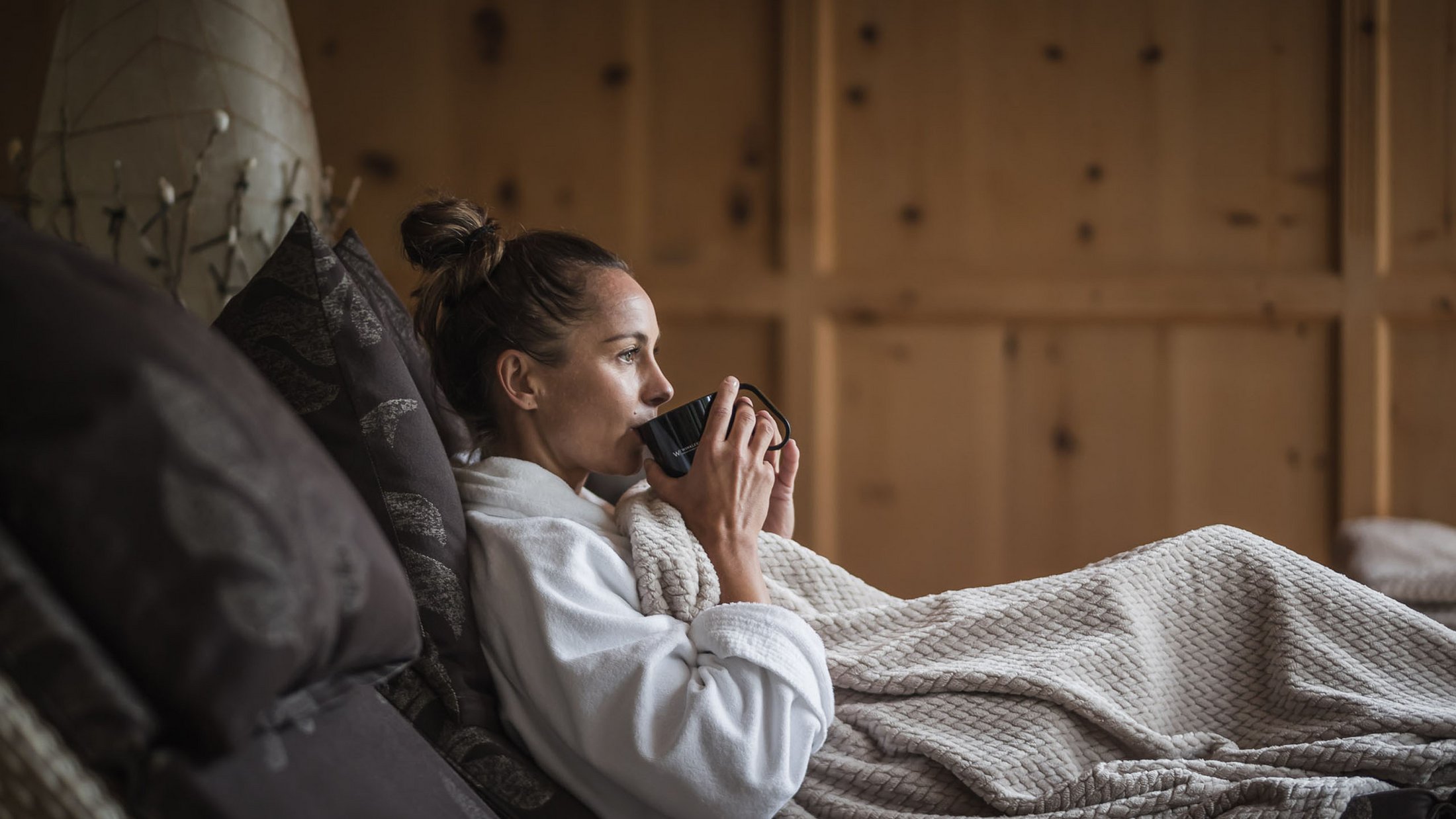 Donna in accappatoio che beve caffè su divano comodo con coperta