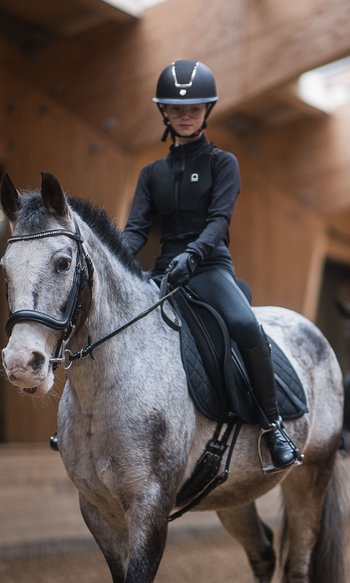 Junge Reiterin auf grauem Pferd in Reithalle mit zweitem Reiter im Hintergrund