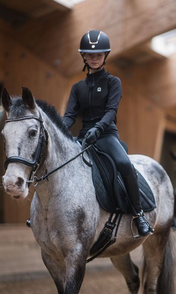 Young rider on gray horse in indoor arena with second rider blurred in background