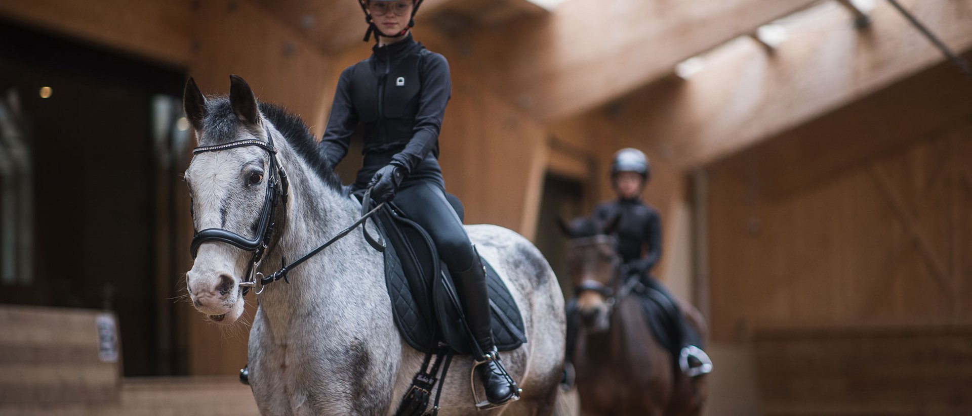 Junge Reiterin auf grauem Pferd in Reithalle mit zweitem Reiter im Hintergrund