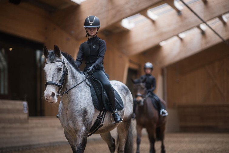 Young rider on gray horse in indoor arena with second rider blurred in background