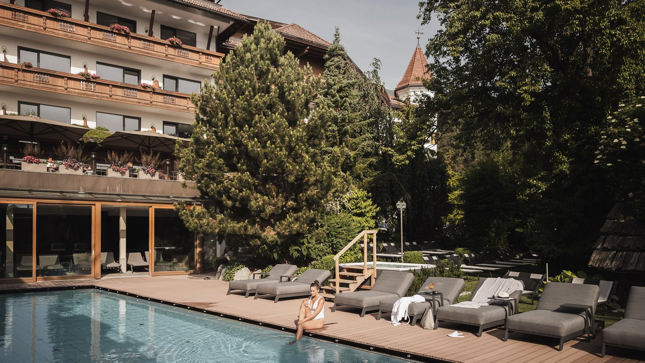 Woman sitting by a hotel pool with lounge chairs and trees in the background