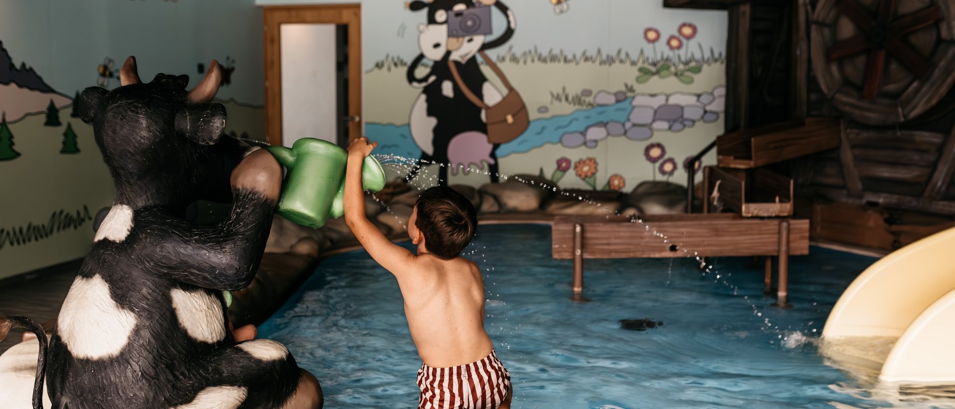 Your source of enrichment: our hotel at Mt Plan de Corones Child playing with watering can near cow statue in indoor pool with mural walls