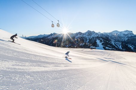 Two skiers on groomed slope with cable cars and mountains at sunset