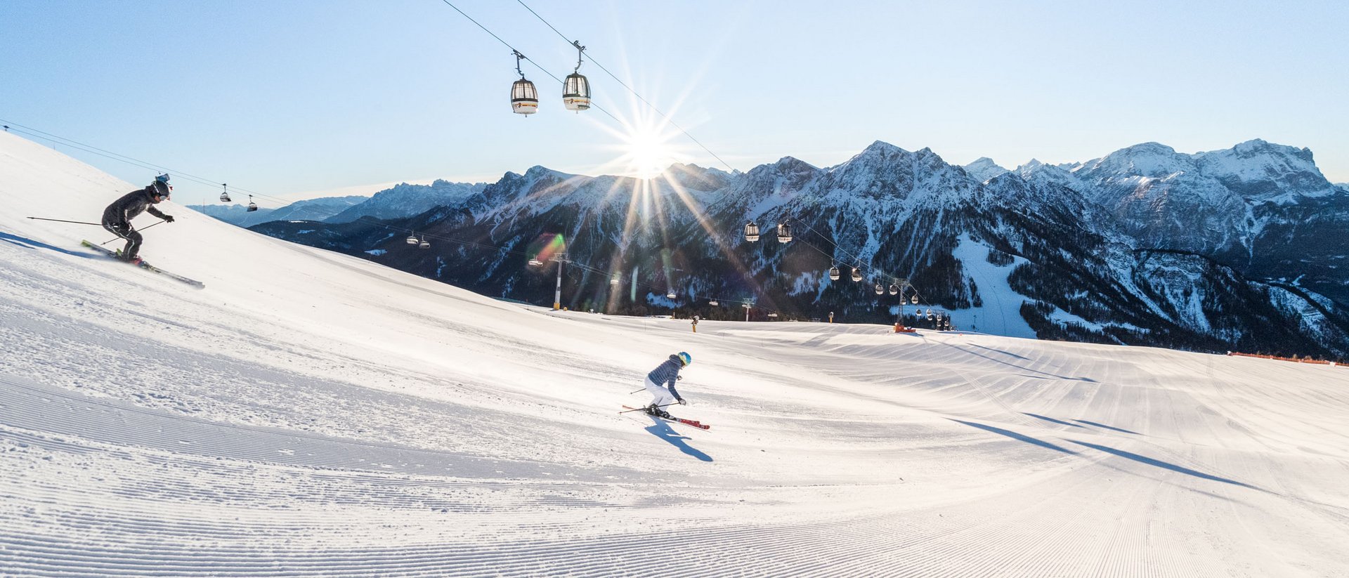 Two skiers on groomed slope with cable cars and mountains at sunset