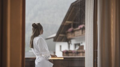 Frau im Bademantel blickt vom Balkon auf ein Nachbarhaus mit Blumen