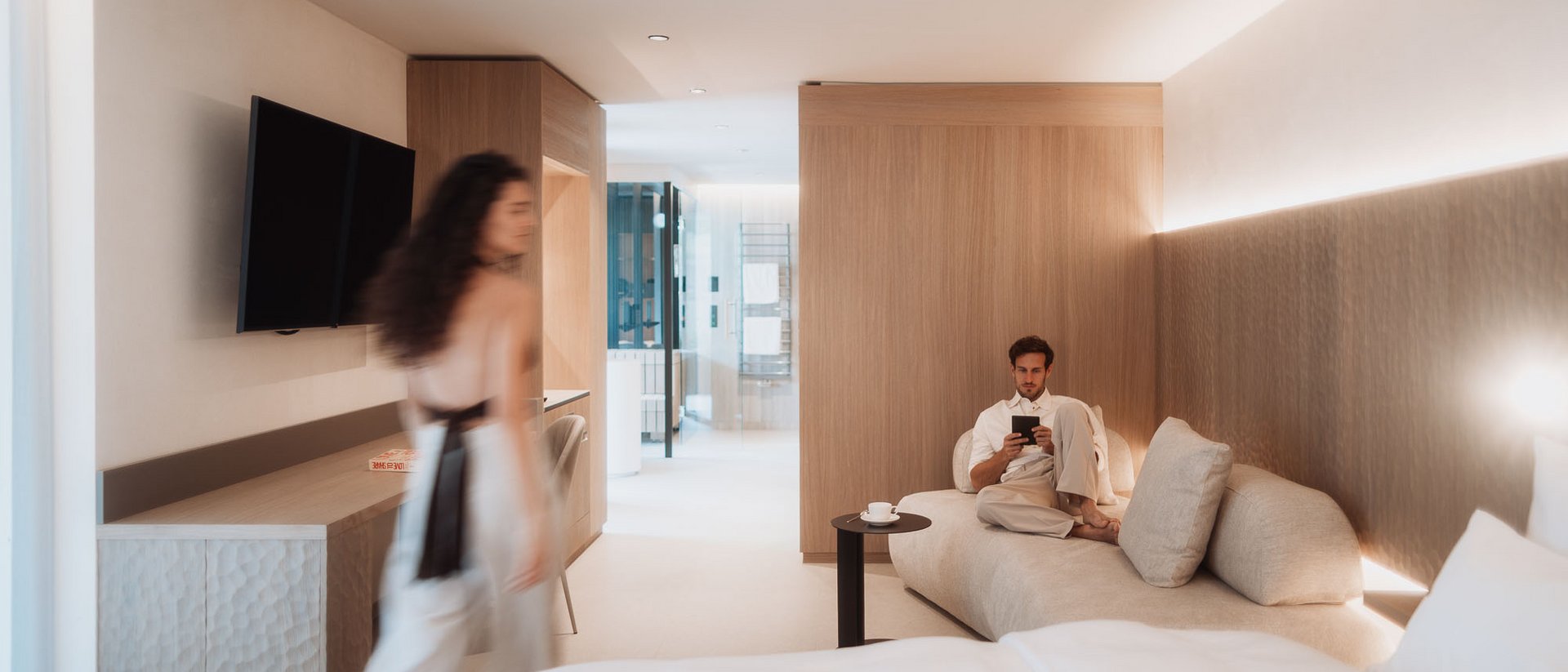 Angolo di pace: il nostro hotel con maneggio in Val Pusteria Uomo seduto sul divano che legge, donna che cammina in una camera d'hotel moderna
