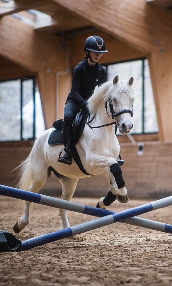 I vostri hotel in Alto Adige: qui dove tutto è possibile Persona cavalca un cavallo bianco saltando un ostacolo in un maneggio