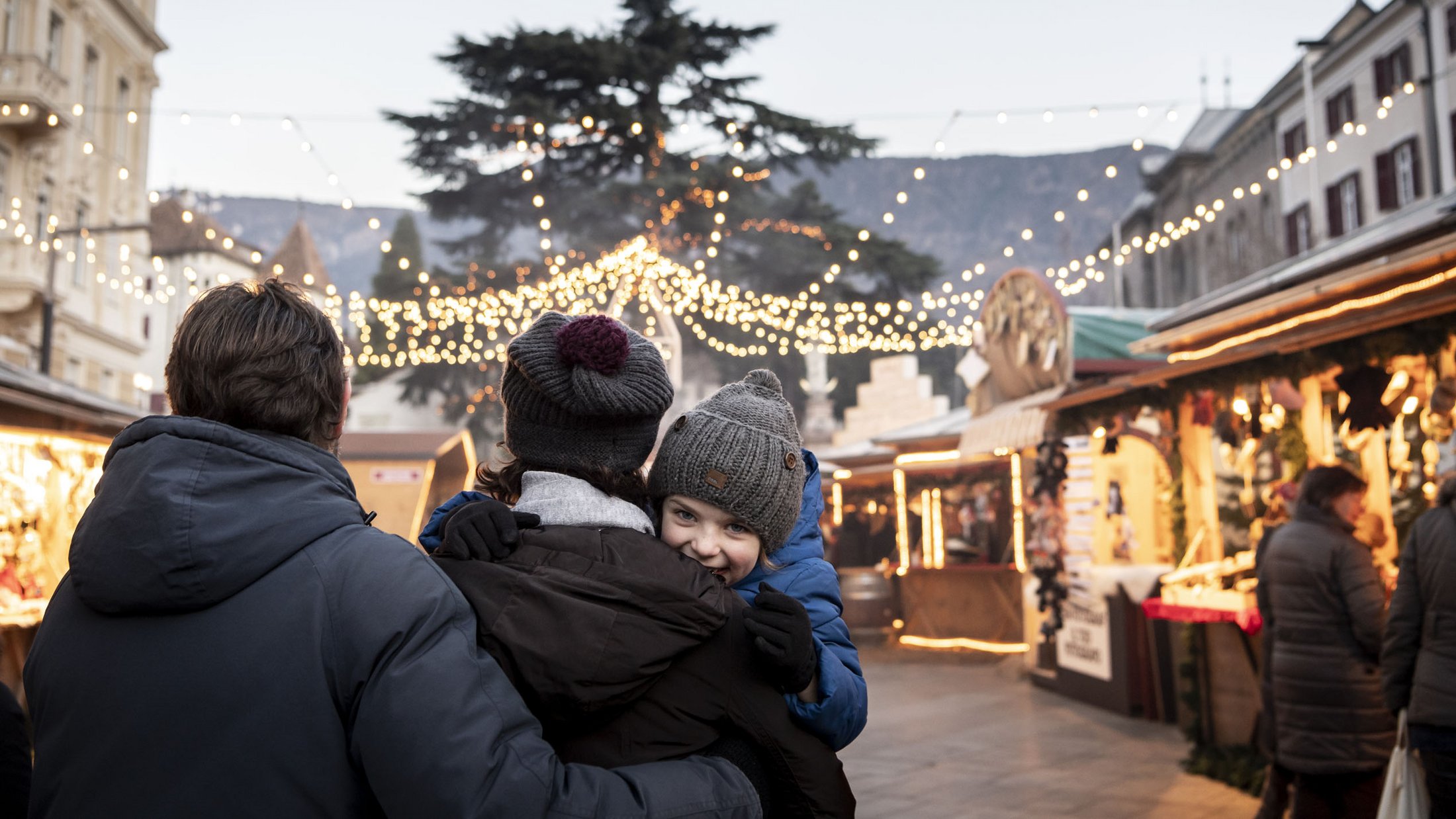Familie beim Weihnachtsmarkt mit Lichterketten und Ständen in der Abenddämmerung