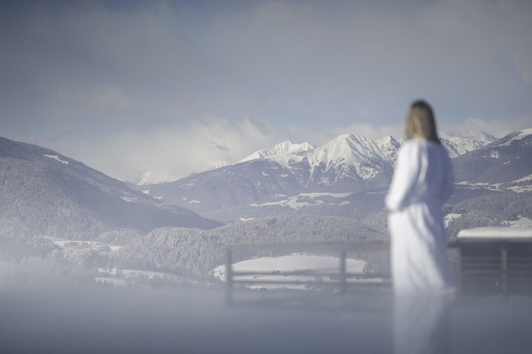 Frau im Bademantel blickt auf schneebedeckte Berge und Wälder