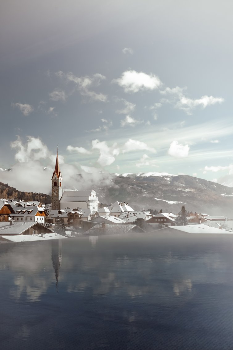 Verschneites Dorf mit Kirche und Bergen im Hintergrund an einem bewölkten Wintertag
